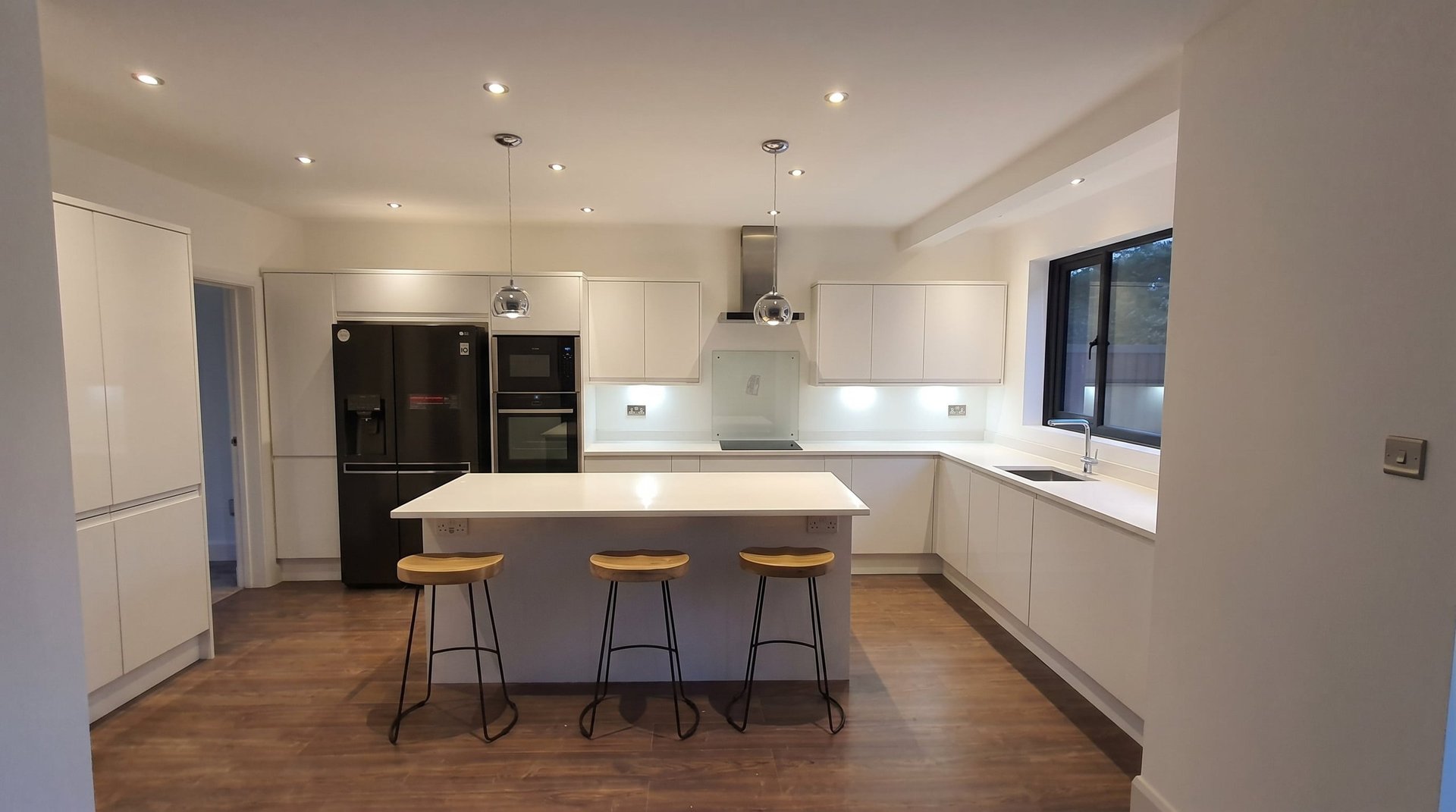 Modern kitchen with white cabinetry, dark countertop island with three bar stools, stainless steel appliances, and wooden flooring.