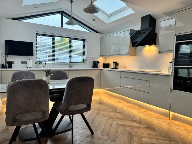 Modern kitchen with white cabinetry, black appliances, island with under-lighting, and two upholstered chairs, bright natural light from skylight