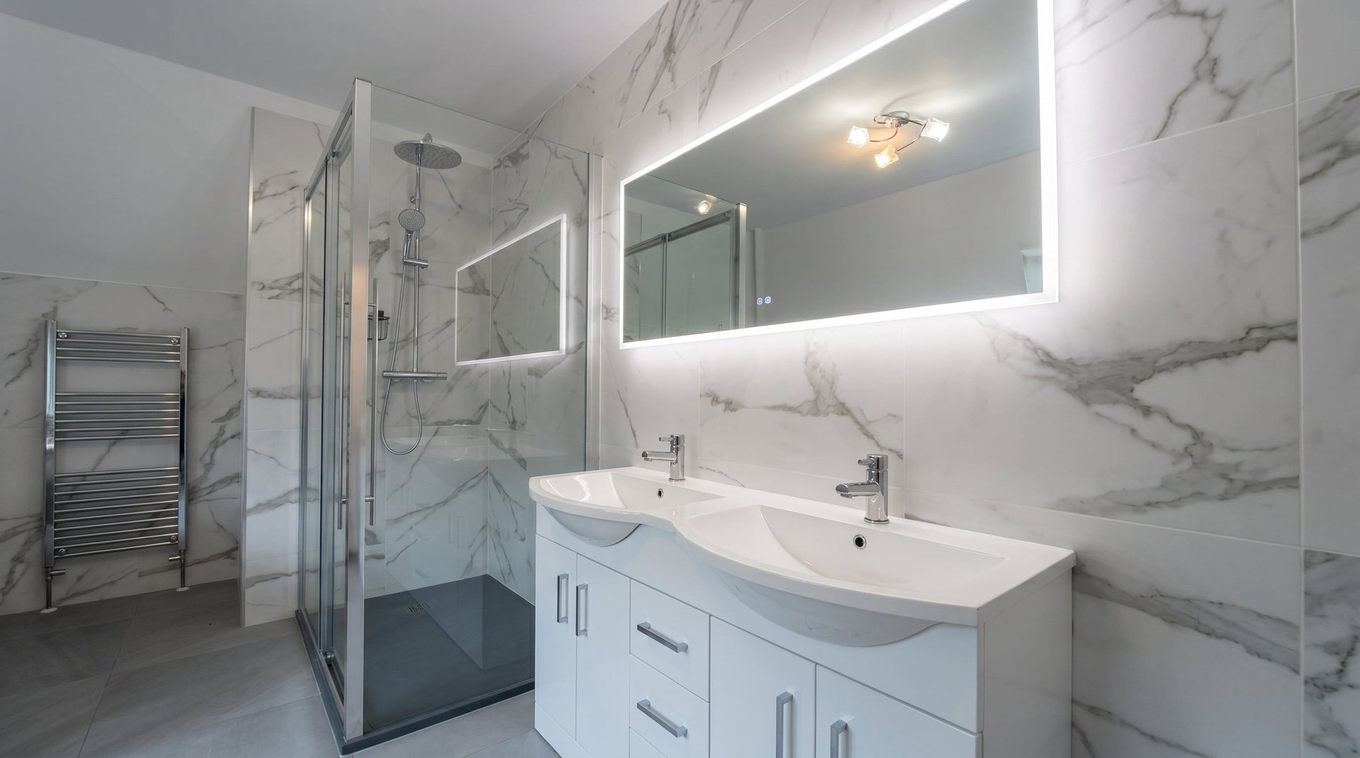 Modern bathroom with marble tile walls, double white vanity sink, illuminated mirror, and glass walk-in shower
