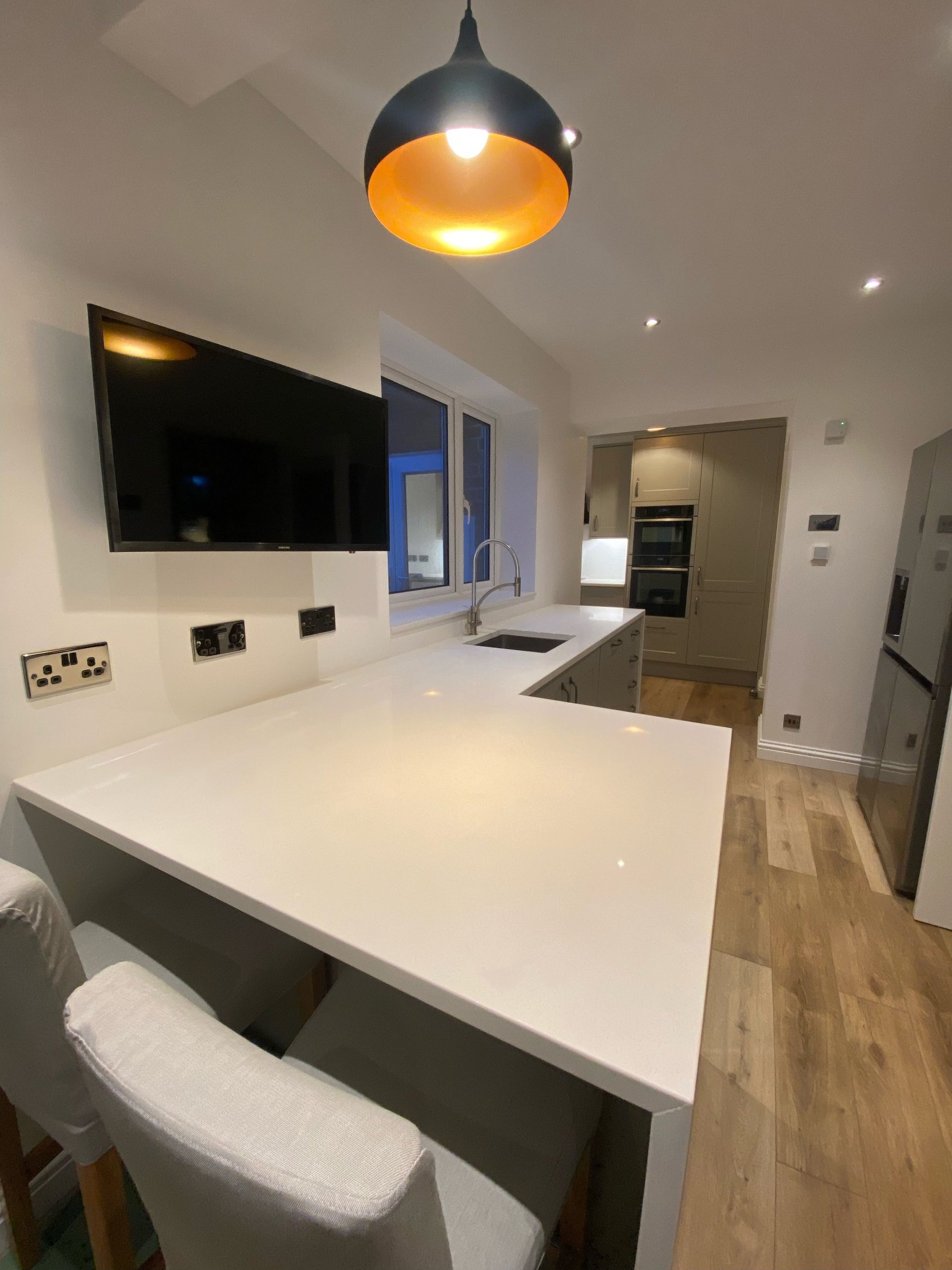 Modern kitchen with white island countertop, black pendant lamp, wall-mounted TV, and stainless steel appliances