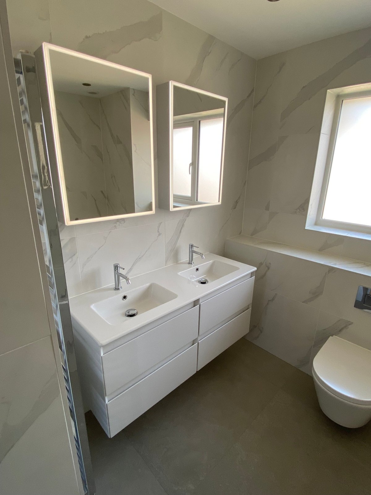 Modern bathroom with white double vanity, illuminated mirrors, marble tile walls, and contemporary white toilet