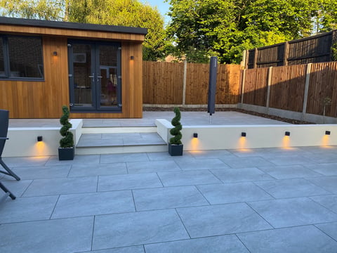 Modern garden patio with blue-grey paving stones, illuminated by pathway lights, featuring a contemporary wooden garden studio with stepped entrance